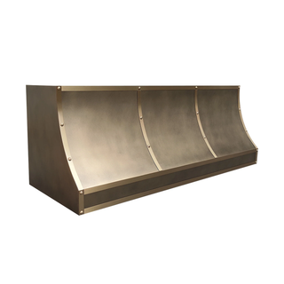 Left angled view of an aged brass vent hood: Elongated profile, curved bottom edges; brushed brass straps with round rivets.