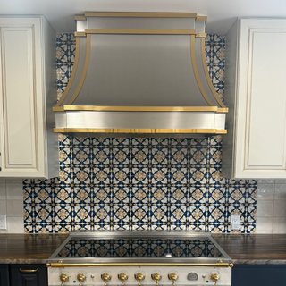 A luxury kitchen stainless steel range hood and freestanding gas stove set against a decorative blue, white, and gold patterned tile backsplash. 