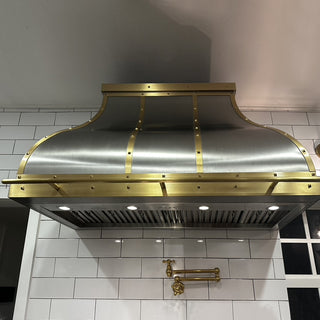 a large stainless steel range hood with brass trim and rivets, mounted above a white subway tile kitchen wall