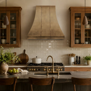 a rustic kitchen scene featuring a large aged brass range hood mounted above a gas stove