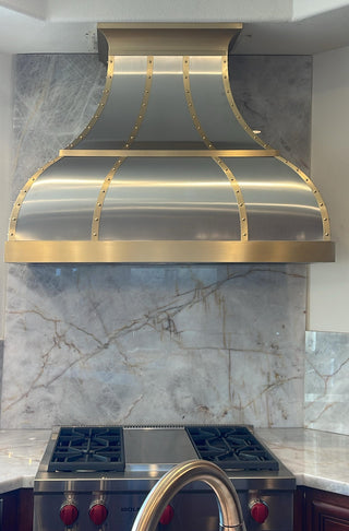 A modern stainless steel range hood with gold trim mounted above a gas stove in a kitchen with a marble backsplash